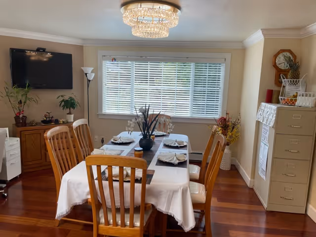 Dining room with a set table for six, wooden chairs, a chandelier, wall-mounted TV, and a window with blinds.