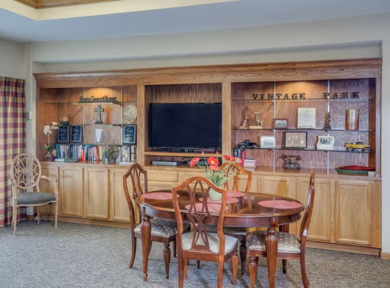 A cozy living room area with a round wooden table surrounded by four chairs with cushioned seats. Behind the table is a built-in wooden shelving unit with a flat-screen TV in the center, books, decorative items, and plaques. The shelves have the words 'Home Sweet Home' and 'Vintage Park' displayed. A single chair is placed to the left near a window with plaid curtains.