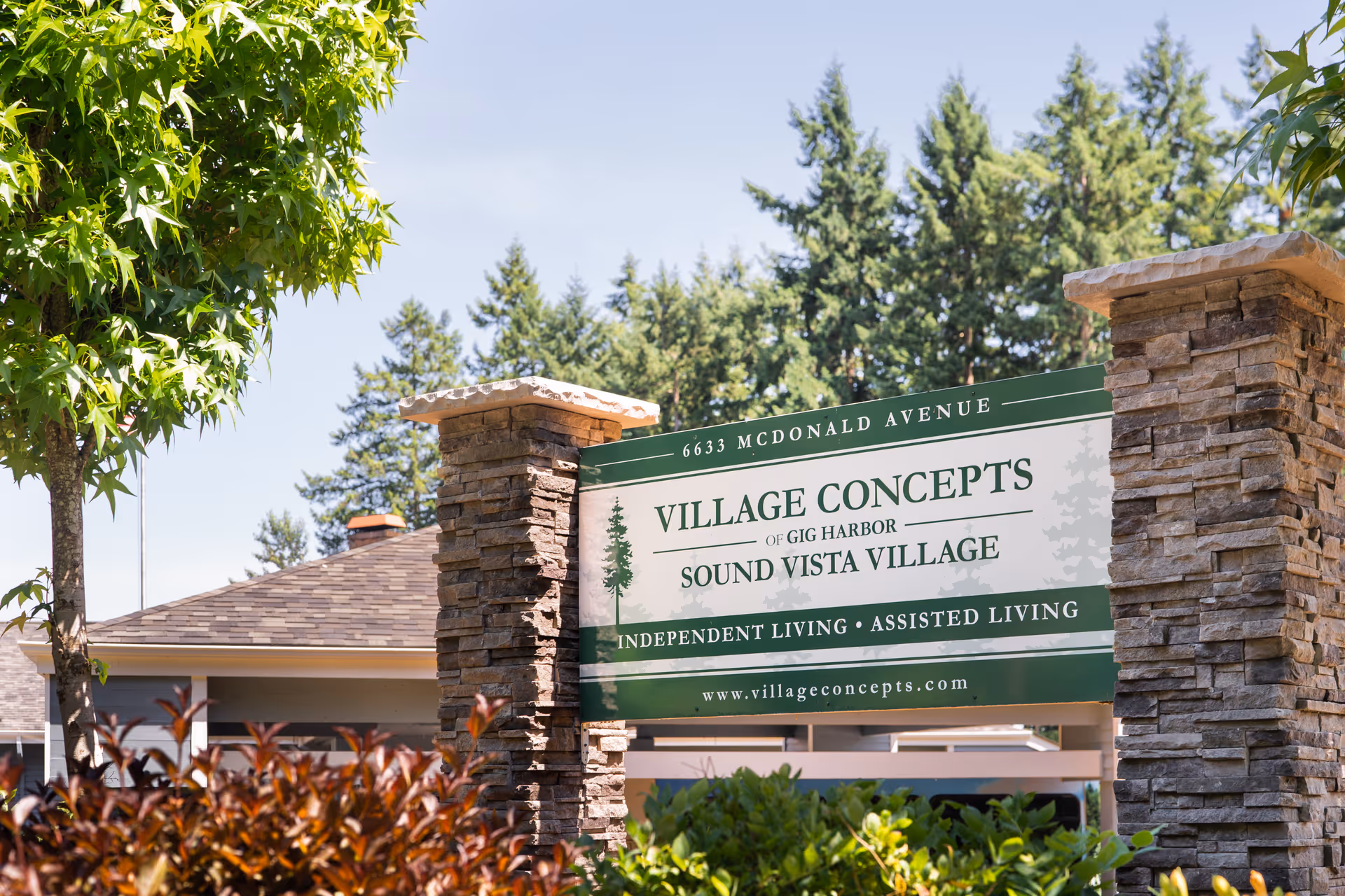 Outdoor image of a stone and wood sign for Village Concepts of Gig Harbor - Sound Vista Village, surrounded by greenery and trees under a clear sky.