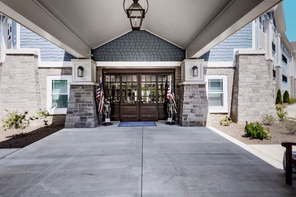 Covered entrance to a building with stone pillars and blue siding, featuring double glass doors flanked by two statues holding American flags and a hanging lantern above.