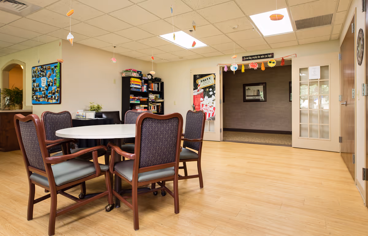 A common area in an assisted living facility with a round white table surrounded by six wooden chairs with padded seats and backs. The room has light wood flooring and a drop ceiling with fluorescent lights. Hanging from the ceiling are small decorations shaped like ice cream cones, hot dogs, and other food items. In the background, there is a black bookshelf filled with board games and books, a blue bulletin board with photos, and a doorway with glass-paneled double doors. Above the doorway is a sign that reads, 'Do one thing everyday that makes you happy.'