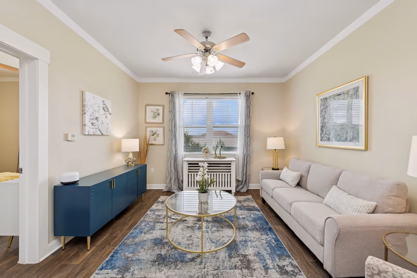 A bright living room with a beige sofa adorned with three cushions on the right, a round glass coffee table with a small potted plant in the center, and a blue sideboard with a lamp and decorative items on the left. The room has light beige walls, a ceiling fan with lights, a window with sheer curtains, and framed artwork on the walls. The floor is wooden with a blue and gray patterned rug.