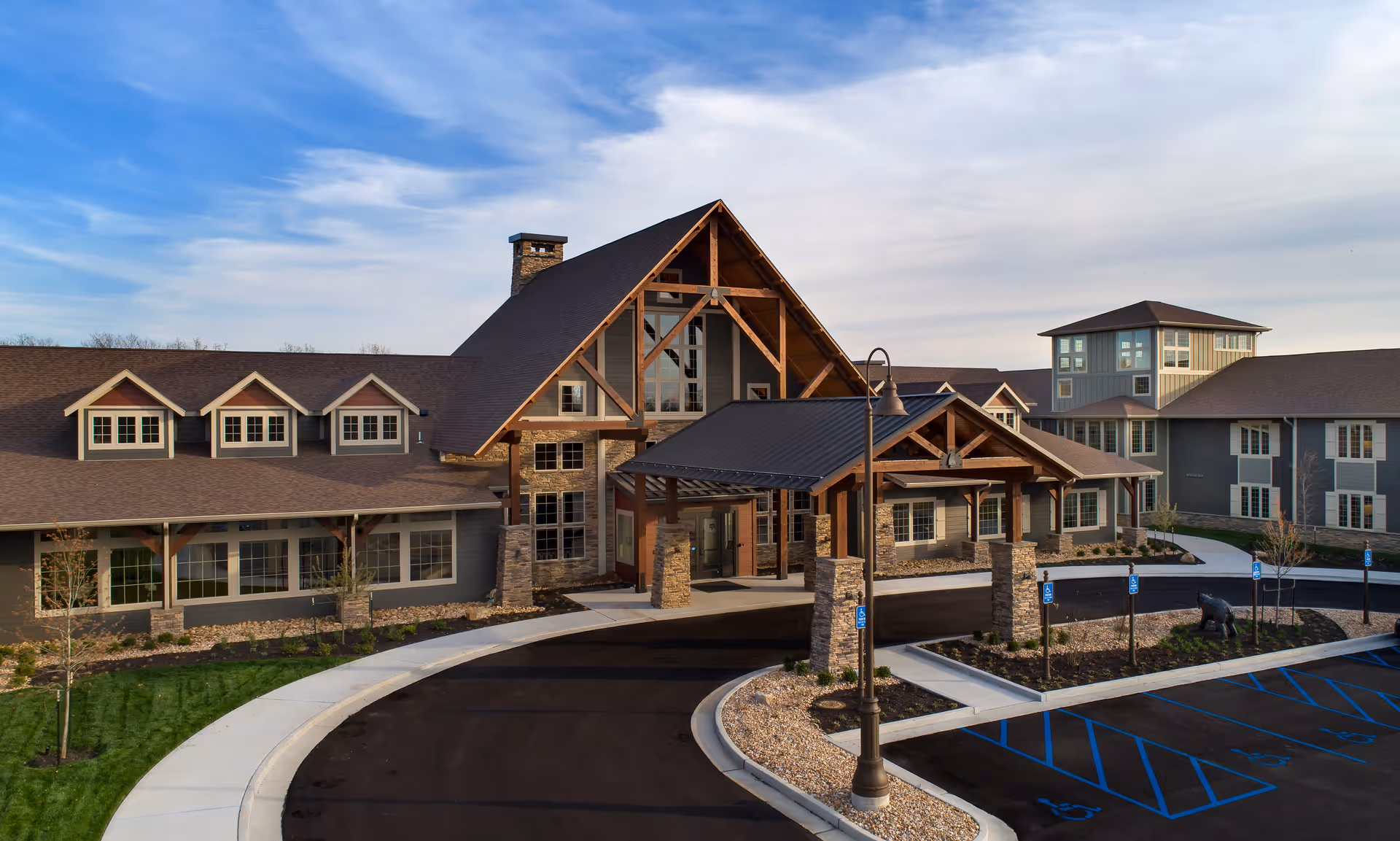 Exterior view of Arrowhead Senior Living facility showing a large building with a peaked roof, stone and wood accents, a covered entrance, and a curved driveway with parking spaces including handicapped spots. The sky is partly cloudy.