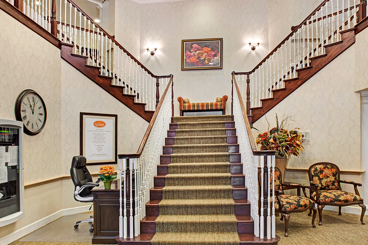 A grand double staircase lobby with carpeted central steps, wooden banisters, seating, floral arrangements and wall art.