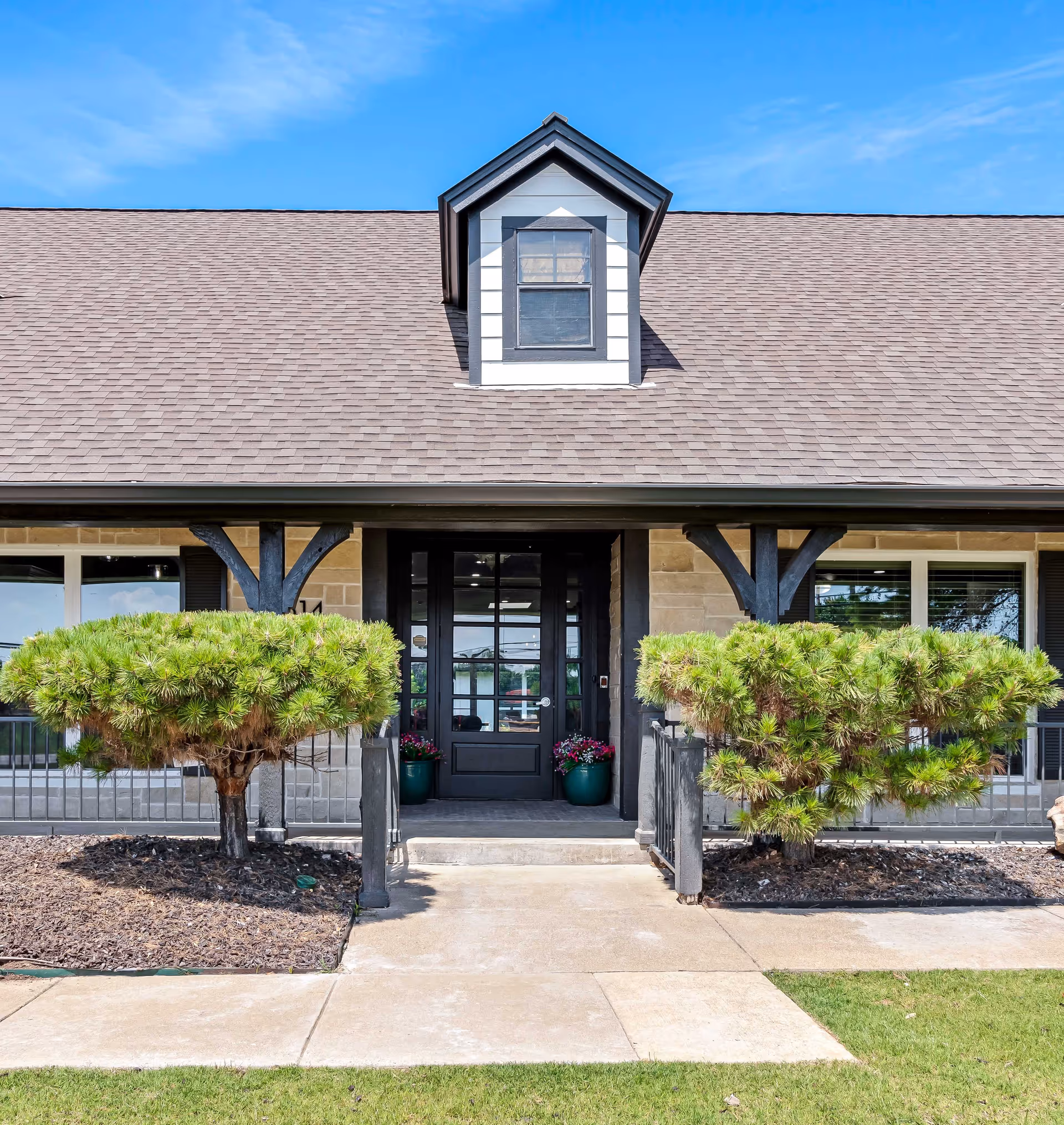 Front entrance of Silver Leaf Assisted Living & Memory Care facility with a black door, two green potted plants with flowers on either side of the door, two trimmed bushes, and a concrete walkway leading to the entrance under a brown shingled roof with a dormer window.