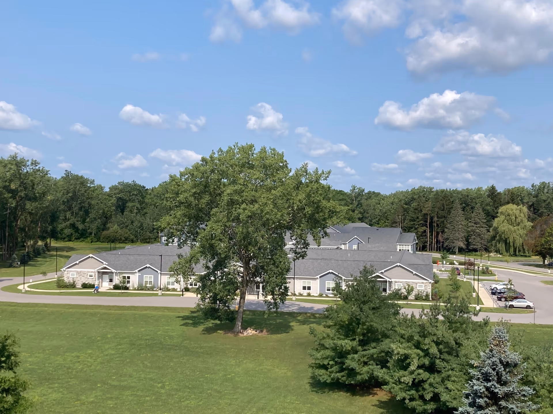 A wide view of the Brothers of Mercy senior living facility building surrounded by green lawns, trees, and a parking area under a partly cloudy blue sky.