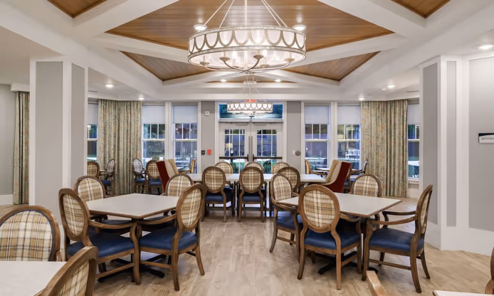 A well-lit dining room with multiple square tables surrounded by wooden chairs with plaid upholstery and blue cushions. The room features large windows with floral curtains, a wooden coffered ceiling with a large circular chandelier, and light-colored wood flooring.