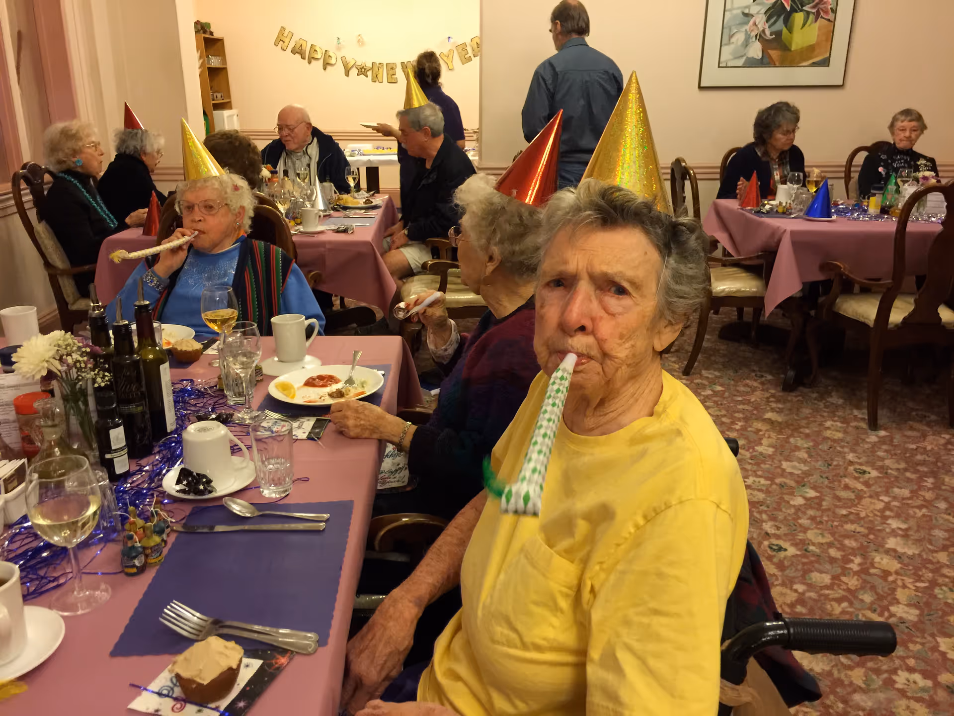 A group of elderly people wearing colorful party hats are seated around tables covered with pink tablecloths, celebrating a New Year's event. Some are blowing party blowers, and there are plates with food, drinks, and decorations on the tables. A 'Happy New Year' banner is visible on the wall in the background.