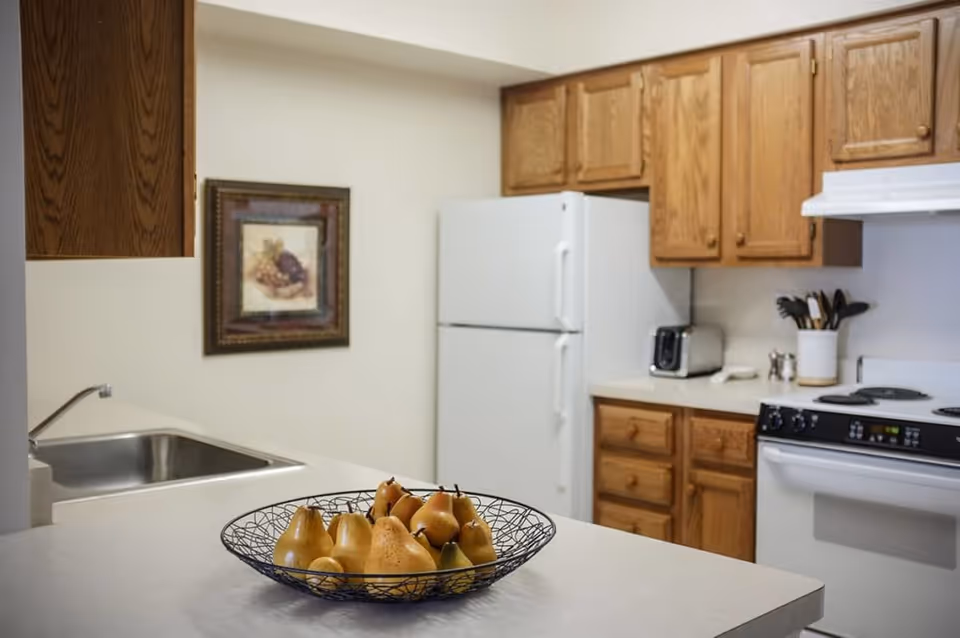 Kitchen with a bowl of pears on a countertop, stainless sink, white refrigerator, wooden cabinets and a stove in the background.