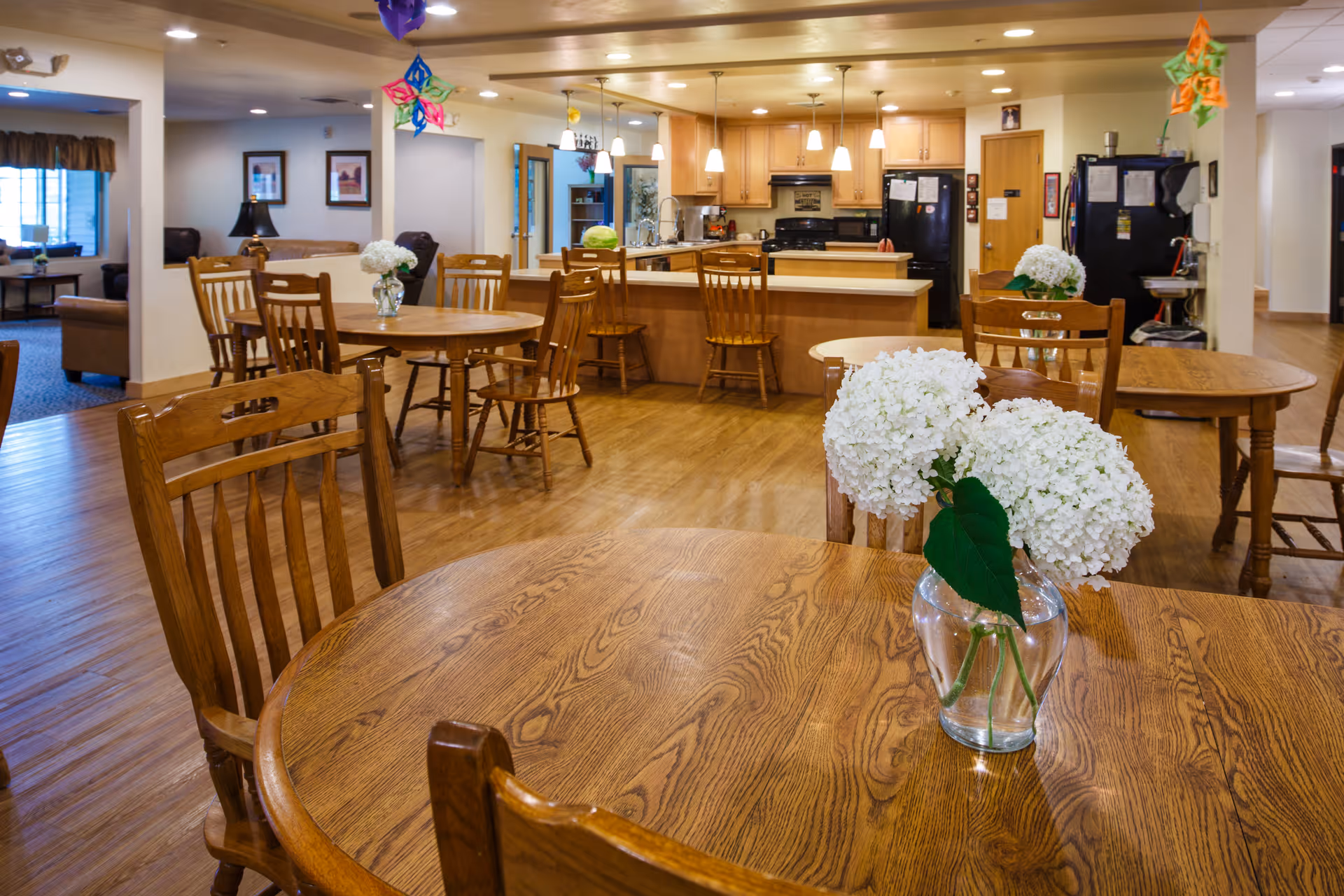 A cozy dining area in a senior living facility featuring multiple wooden tables and chairs. Each table has a vase with white hydrangea flowers. In the background, there is a kitchen with wooden cabinets, a black refrigerator, and pendant lights hanging from the ceiling. The room has wooden flooring and is decorated with colorful hanging ornaments.