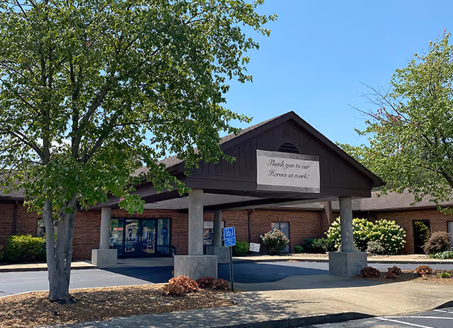 Exterior view of a single-story brick building with a covered entrance supported by concrete pillars. There are trees and landscaping around the entrance. A sign hanging on the building reads 'Thank you to our Heroes at work!'. A handicapped parking sign is visible near the entrance.