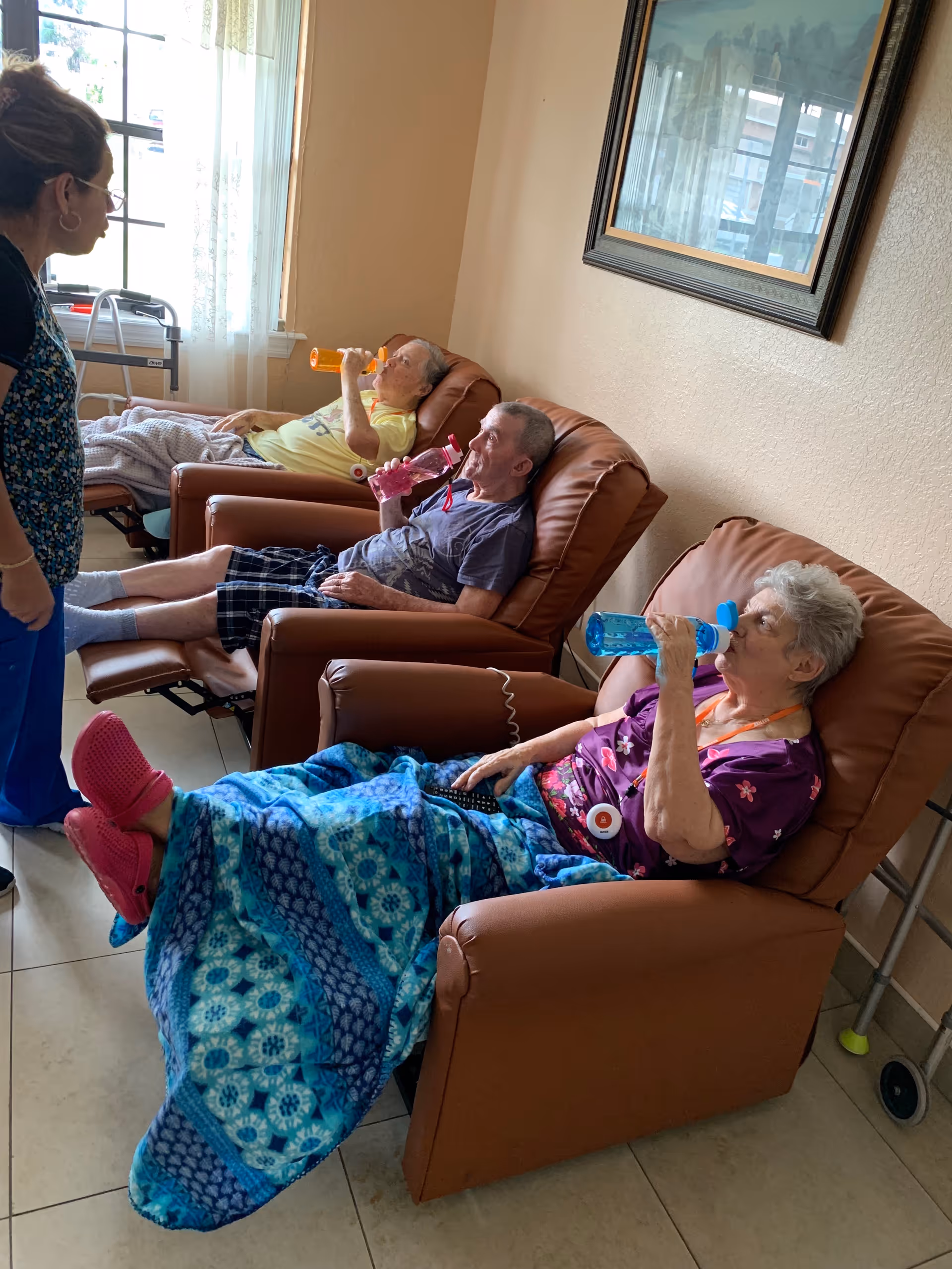 Three elderly individuals sitting in reclining chairs in a room, each drinking from colorful water bottles. A caregiver stands nearby, engaging with them. One person is covered with a blue patterned blanket and wearing pink slippers. There is a window with sheer curtains in the background and a framed picture on the wall.