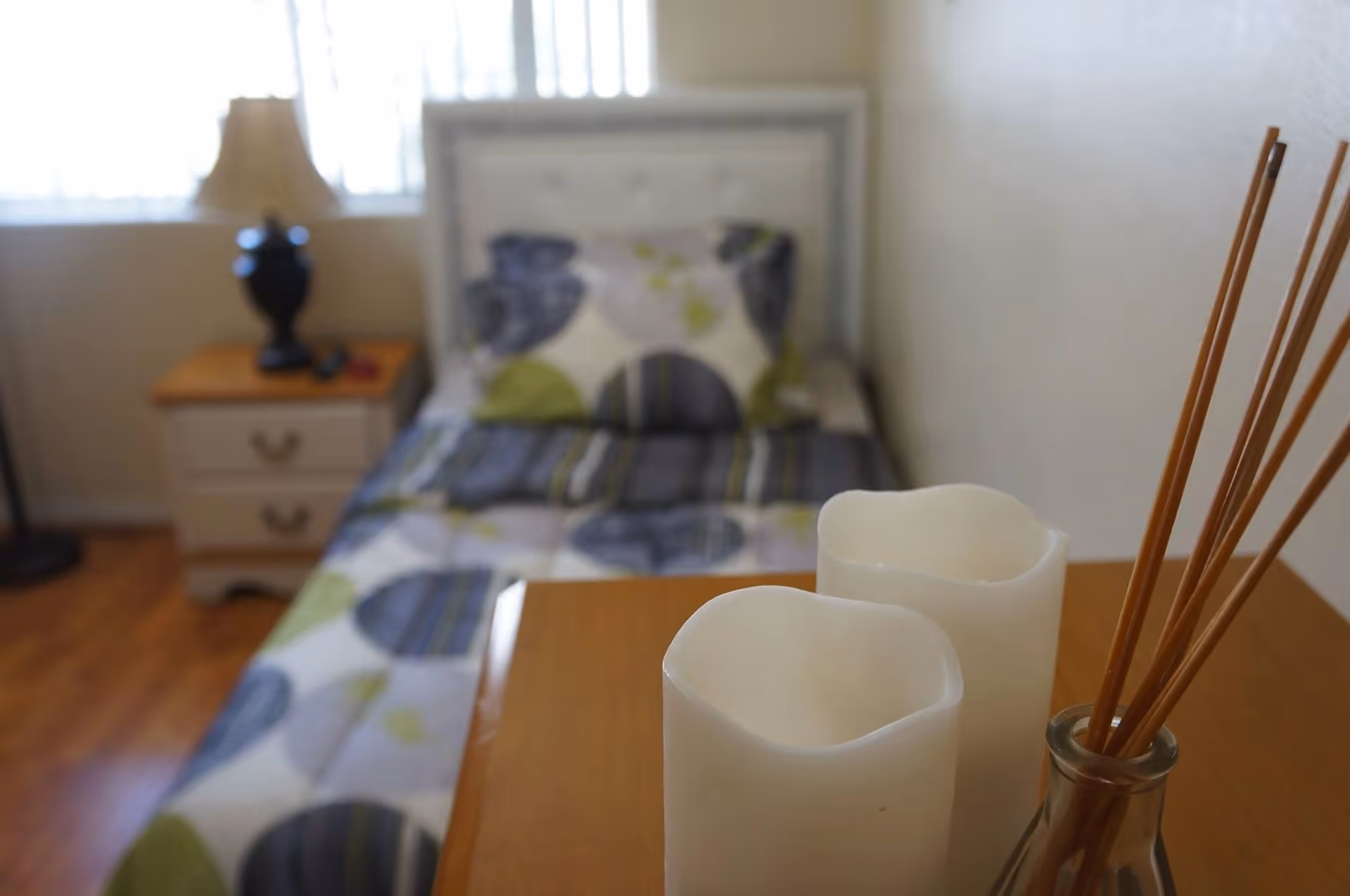 A cozy bedroom with a single bed covered in a patterned bedspread featuring blue, green, and white colors. Next to the bed is a wooden nightstand with two drawers, a black lamp with a beige lampshade, and some small items. In the foreground, there are two white candles and a glass container holding reed diffuser sticks on a wooden surface.