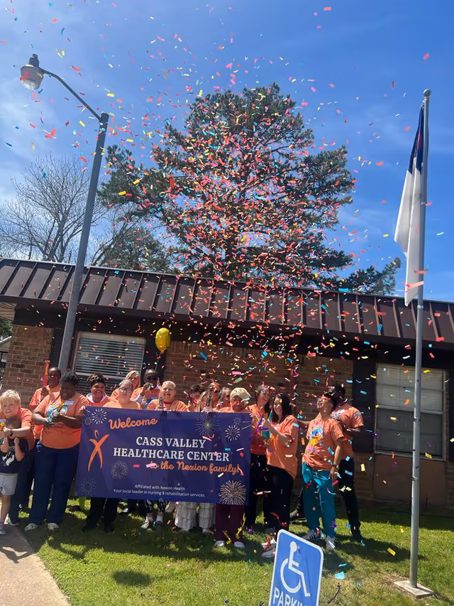 A group of people standing outside a brick building holding a purple banner that reads 'Welcome Cass Valley Healthcare Center the Nexion family.' Colorful confetti is falling from above, and there is a flagpole with a white flag and a blue handicap parking sign in the foreground.