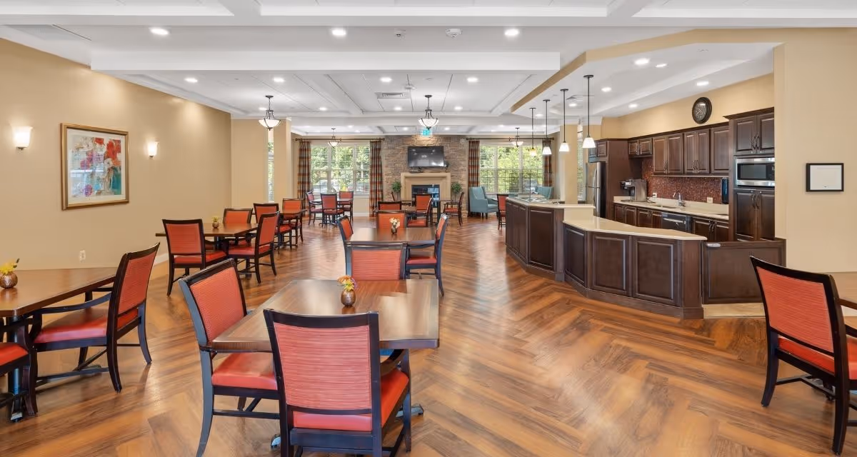 A spacious dining area with multiple wooden tables and red cushioned chairs arranged neatly on a wooden floor. To the right, there is a kitchen area with dark wood cabinets, a countertop, and hanging pendant lights. At the far end of the room, large windows with plaid curtains let in natural light, and a stone fireplace with a mounted TV above it is centered on the back wall.