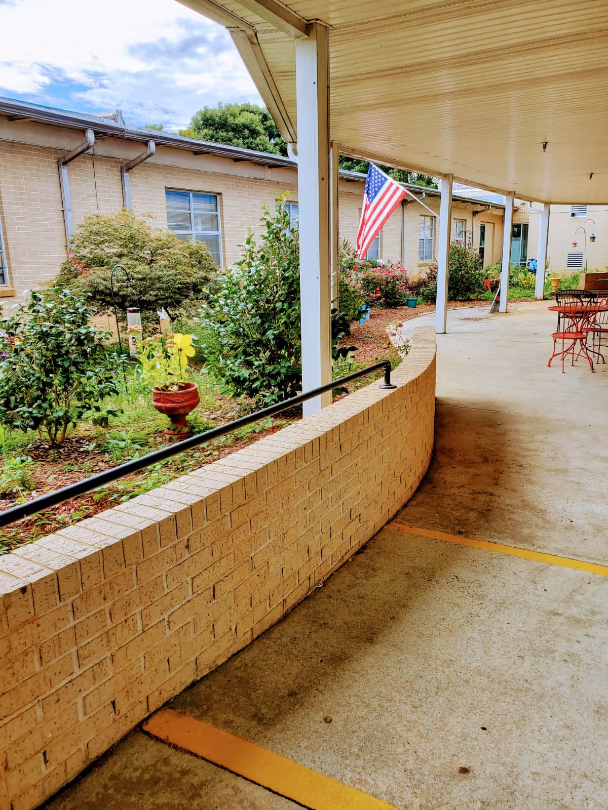 Covered outdoor walkway with a low brick wall and metal railing on the left side, overlooking a garden with bushes, flowers, and an American flag. There are red metal chairs and tables on the right side under the covered area. The building with windows is visible in the background under a partly cloudy sky.