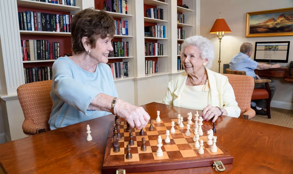 Two elderly women sitting at a wooden table playing chess in a room with bookshelves filled with books behind them. One woman is making a move while both smile at each other. In the background, an elderly man is seated at a desk using a computer. The room is warmly lit with a floor lamp and a framed painting on the wall.