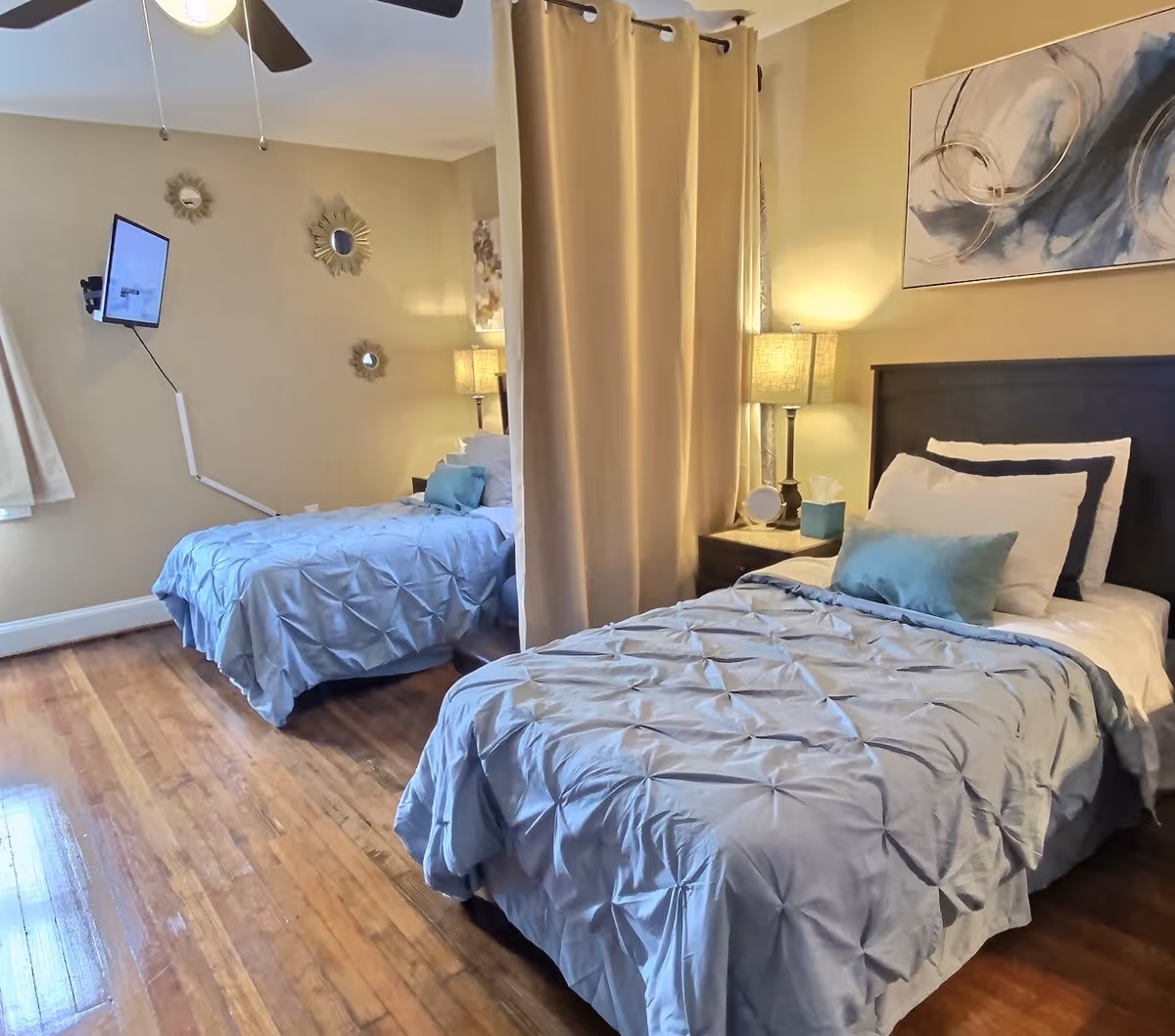 Two twin beds in a warmly lit bedroom with a curtain divider, bedside lamps, wall art, and a mounted TV.