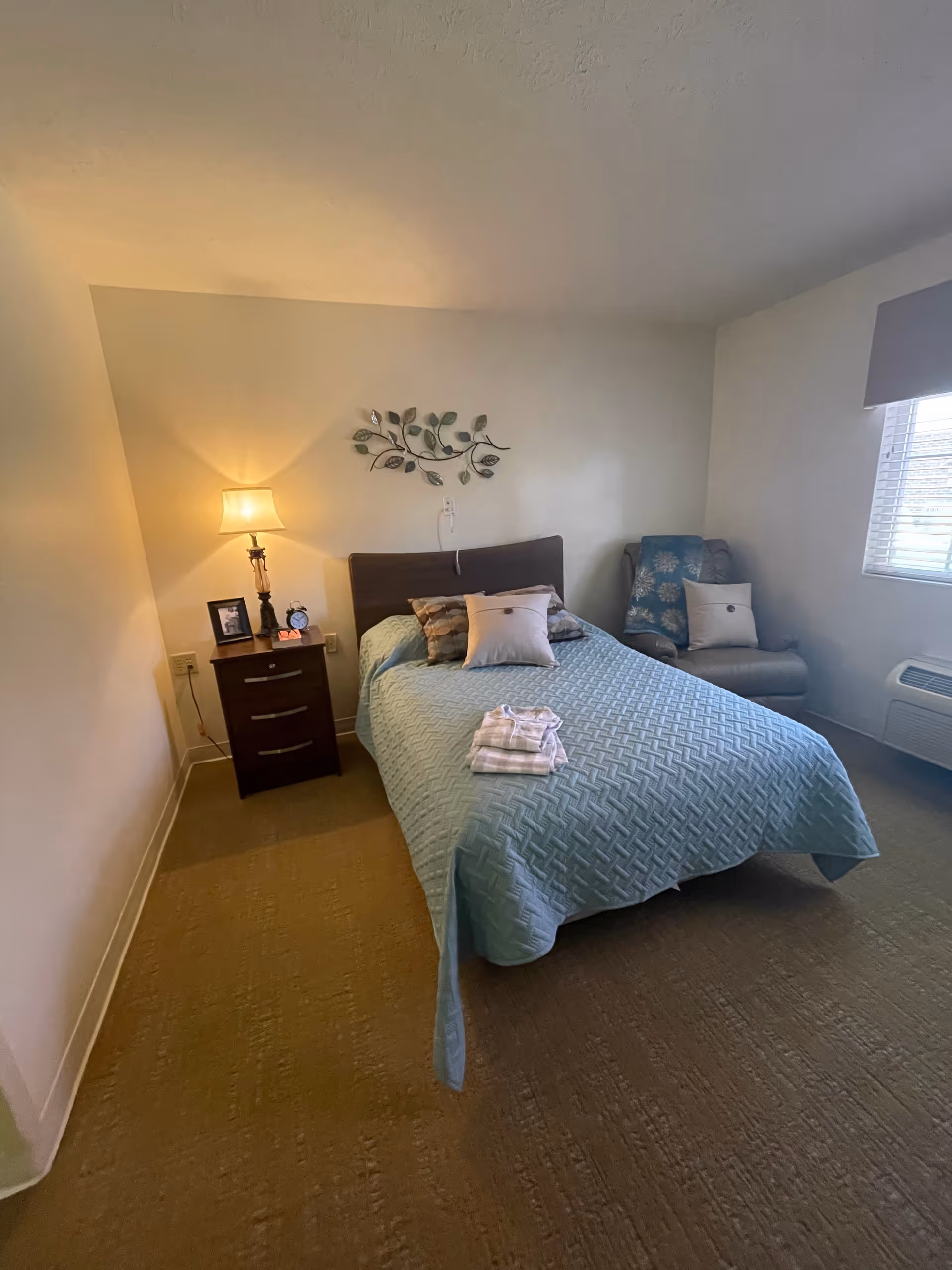 A tidy bedroom with a single bed covered in a blue quilt, a bedside table with a lamp and clock, a decorative wall sculpture above the headboard, and an upholstered chair by a window.