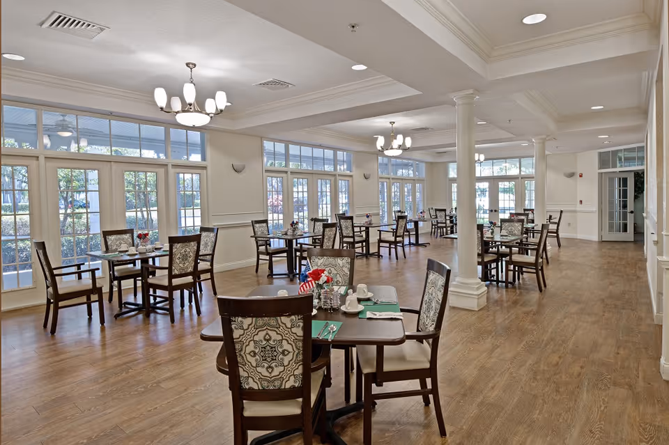 A spacious dining room with multiple wooden tables and chairs arranged neatly. Each table is set with placemats, cups, and utensils. Large windows and glass doors line the walls, allowing natural light to fill the room. The floor is wooden, and the ceiling has recessed lighting and chandeliers. The room has white walls and decorative columns.