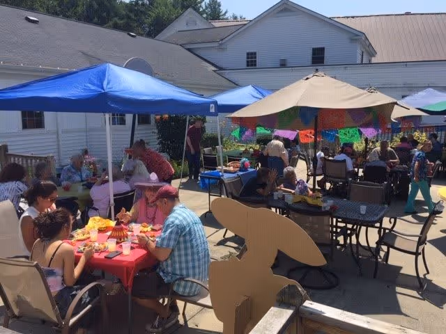 Outdoor patio area at Pine Rock Manor with several people sitting at tables under blue and beige umbrellas, enjoying food and socializing on a sunny day. Colorful decorations hang from the beige umbrella, and the building of the facility is visible in the background.