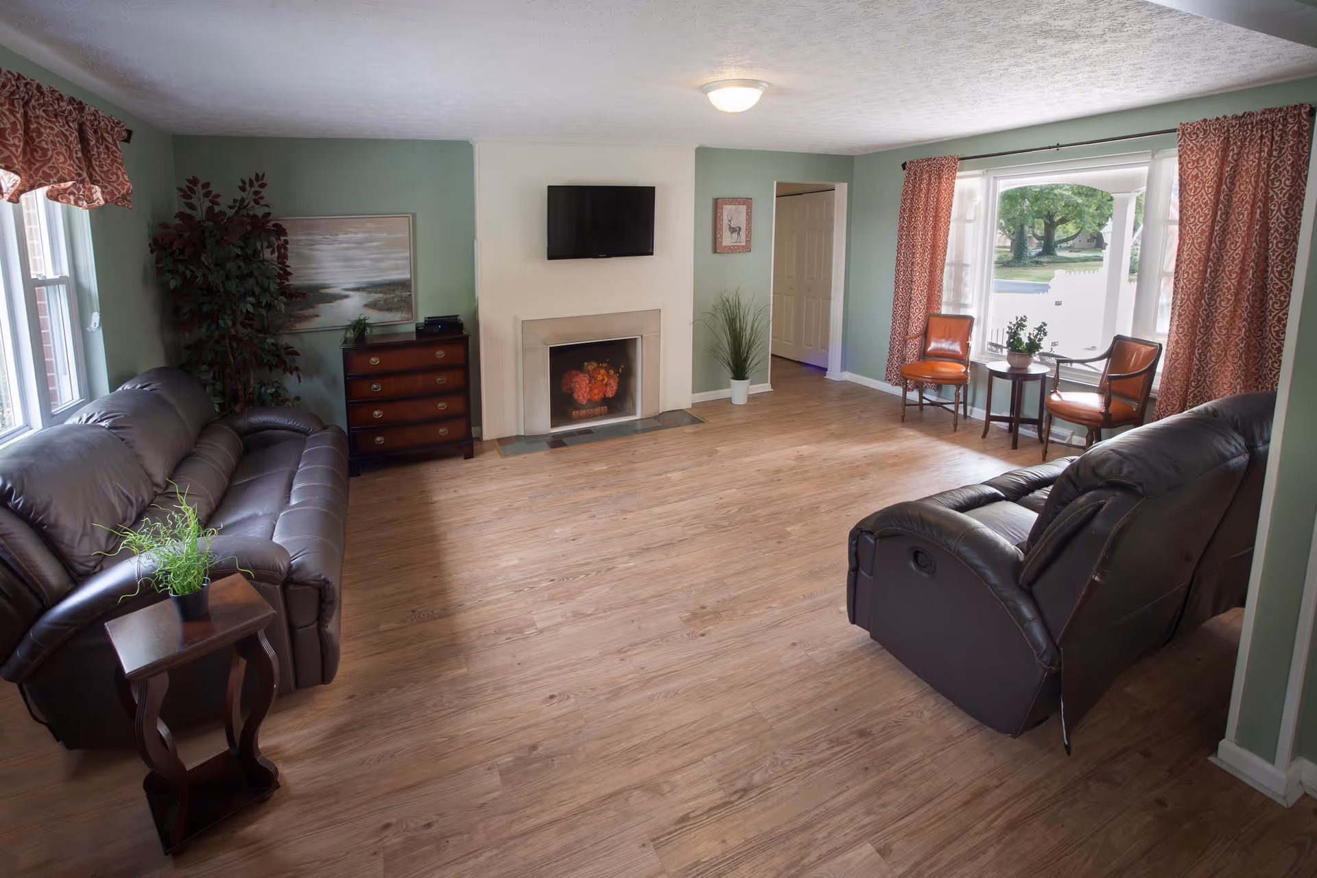 A cozy living room with two dark brown leather sofas facing each other, a small wooden side table with a plant, a wooden chest of drawers, a flat-screen TV mounted above a fireplace with floral decoration, and two brown chairs with a small round table near a large window with red patterned curtains. The room has light wood flooring and green walls.