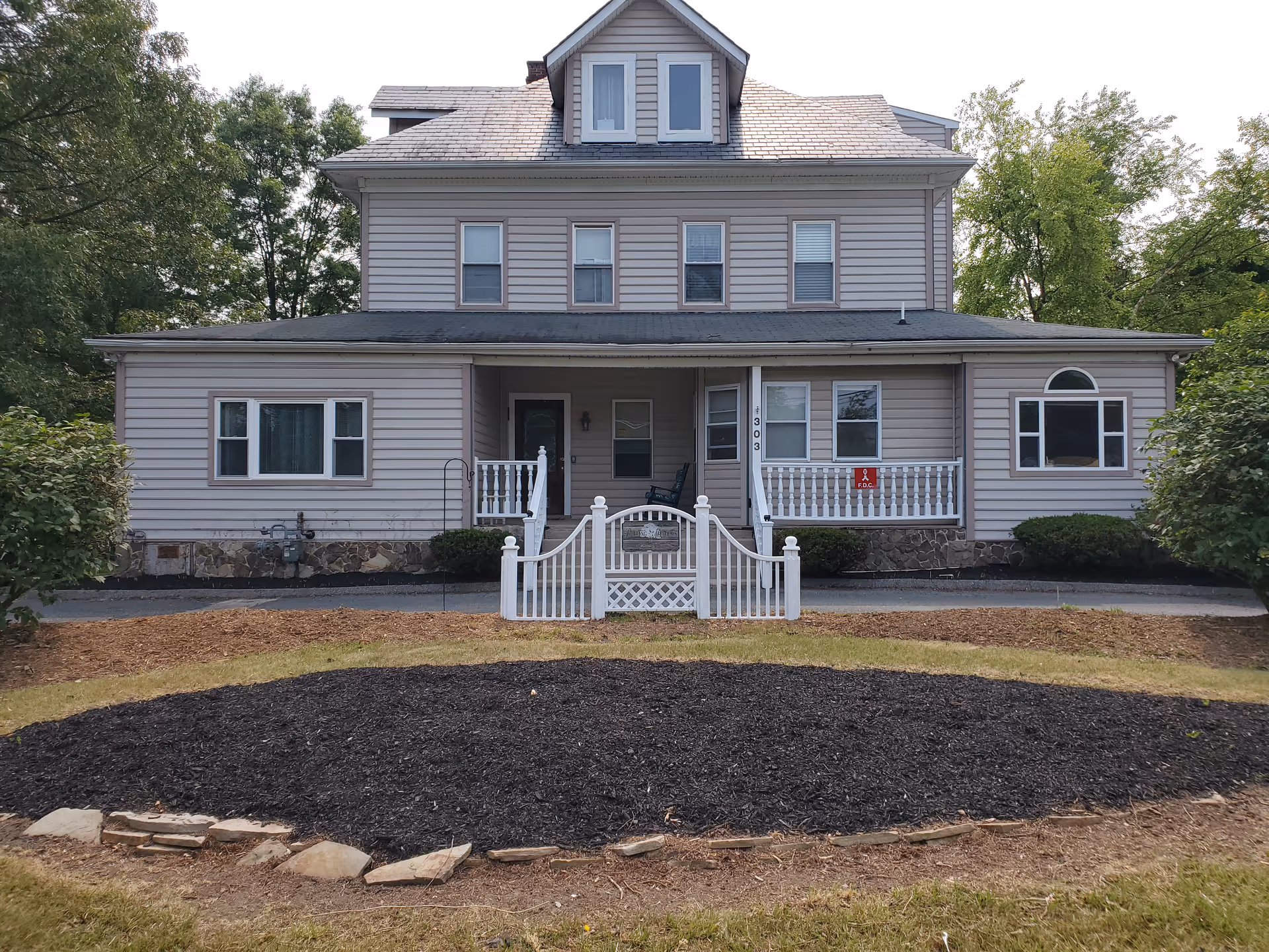 Front exterior of a multi-story beige house with a covered porch, white picket gate, and a landscaped front yard.
