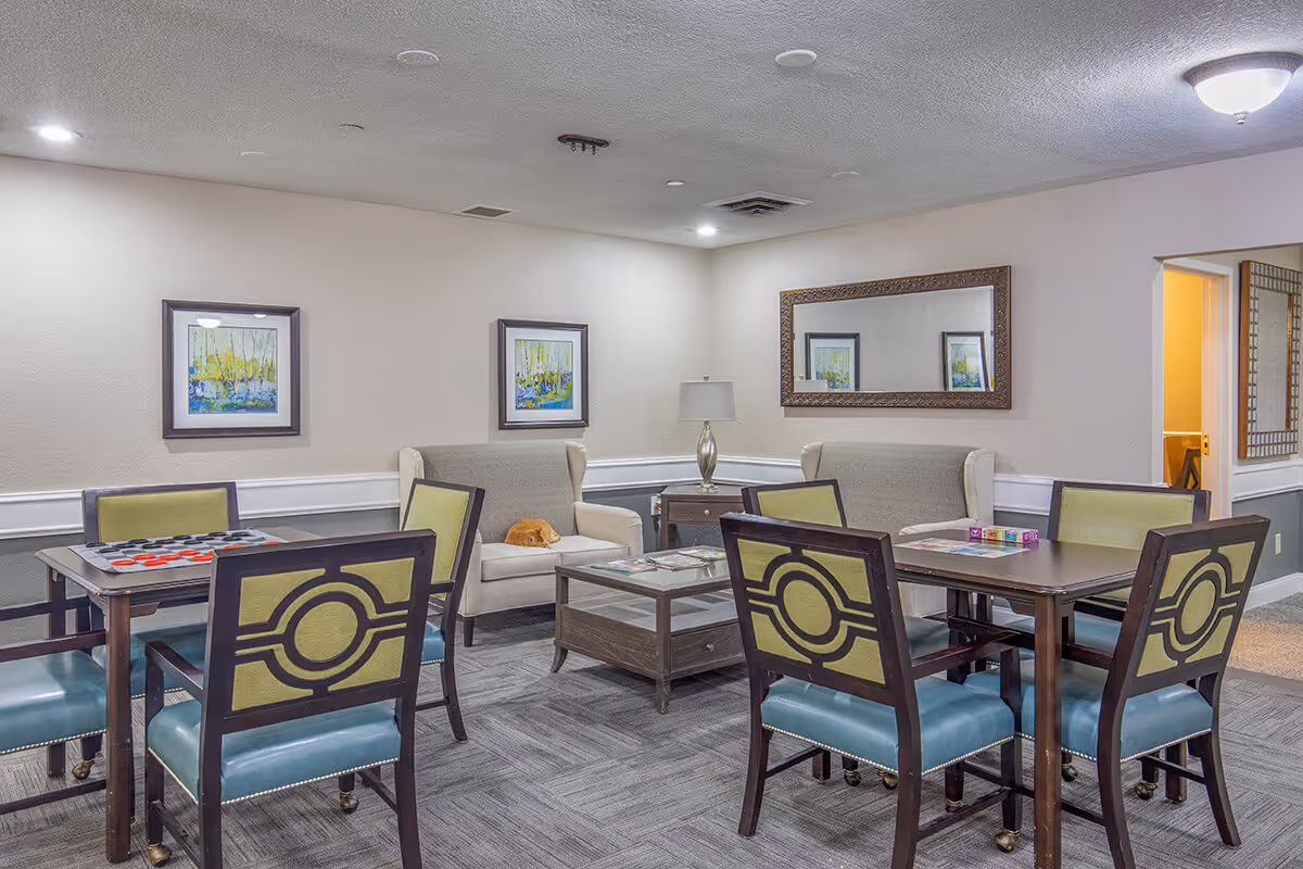 A cozy common area in Arlington Senior Living featuring two tables with green and blue cushioned chairs, two beige armchairs with a small orange cat sleeping on one, a wooden coffee table, a side table with a lamp, two framed paintings on the wall, and a large decorative mirror reflecting part of the room.