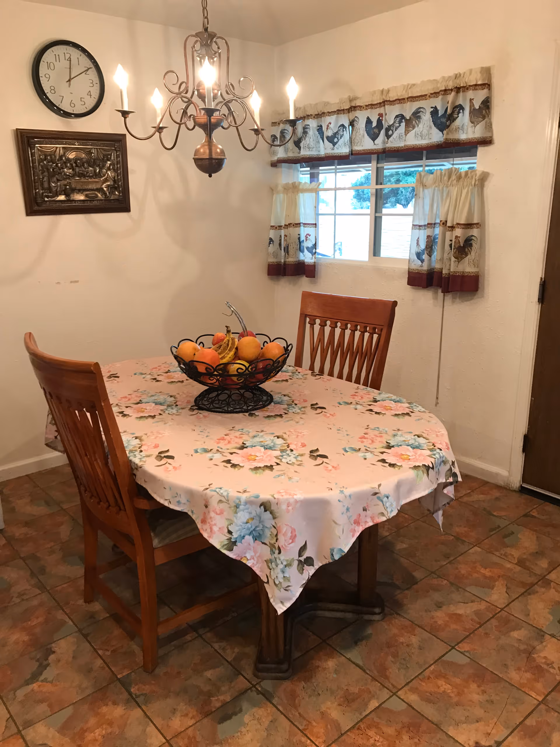 A cozy dining room with a wooden table covered by a floral tablecloth. On the table is a black wire fruit basket filled with various fruits. Two wooden chairs are placed around the table. The room has tiled flooring, a window with curtains featuring a rooster pattern, a wall clock, and a decorative wall plaque. A chandelier with lit bulbs hangs above the table.