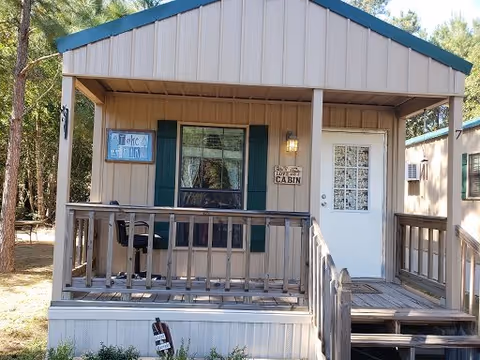 Small cabin with a covered porch featuring a wooden railing, a chair, and a door with a window. The cabin has beige siding with green shutters around the window. There are signs on the cabin that read 'Take it easy' and 'Enjoy your stay at the cabin'. The cabin is surrounded by trees.