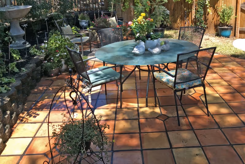 Outdoor patio area with a round glass table surrounded by four metal chairs with cushions. The patio has terracotta tiles and is decorated with potted plants and flowers. A wooden fence and greenery are visible in the background.
