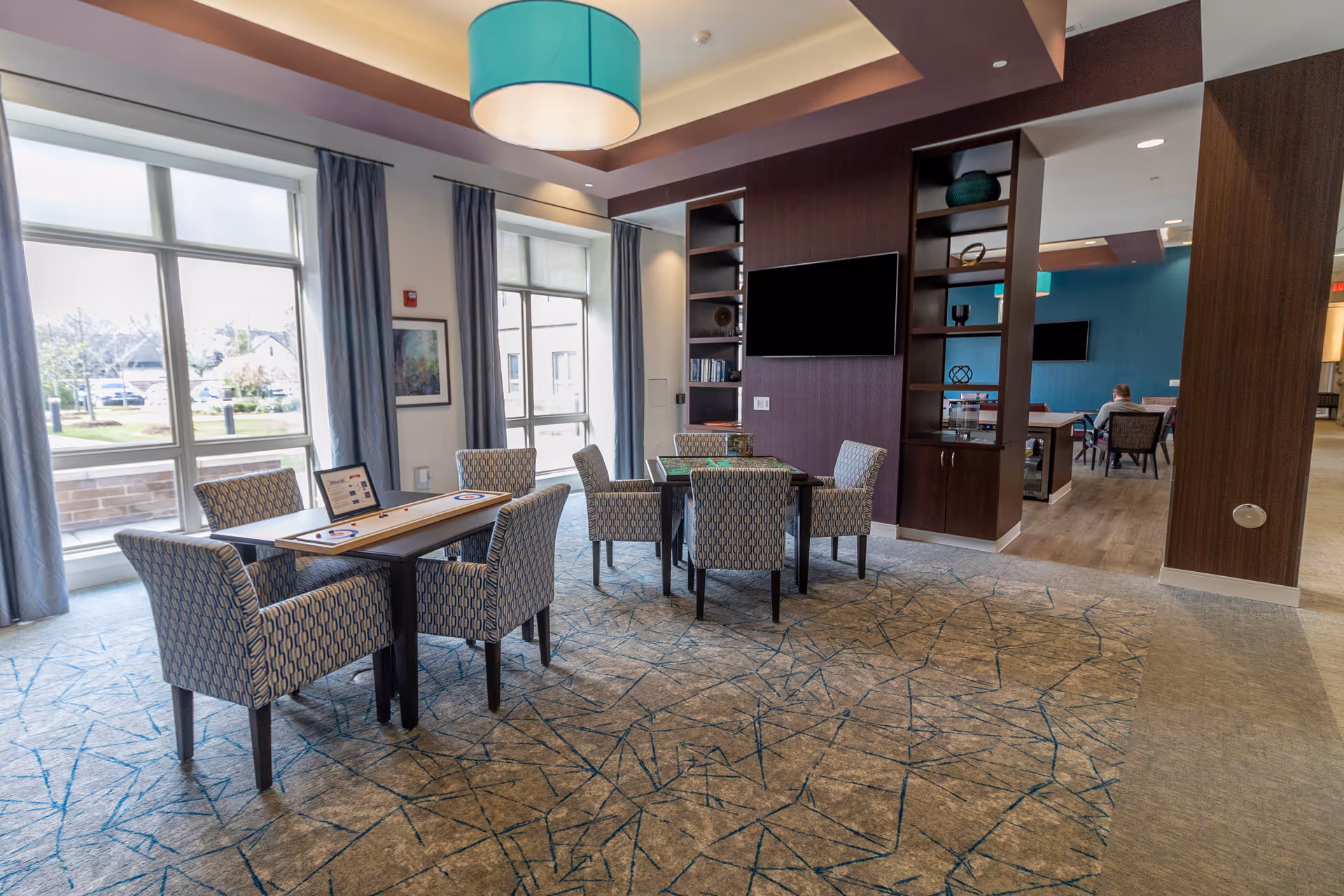 Bright communal seating area with tables, upholstered chairs, built-in shelving and a wall-mounted TV in a senior living facility.