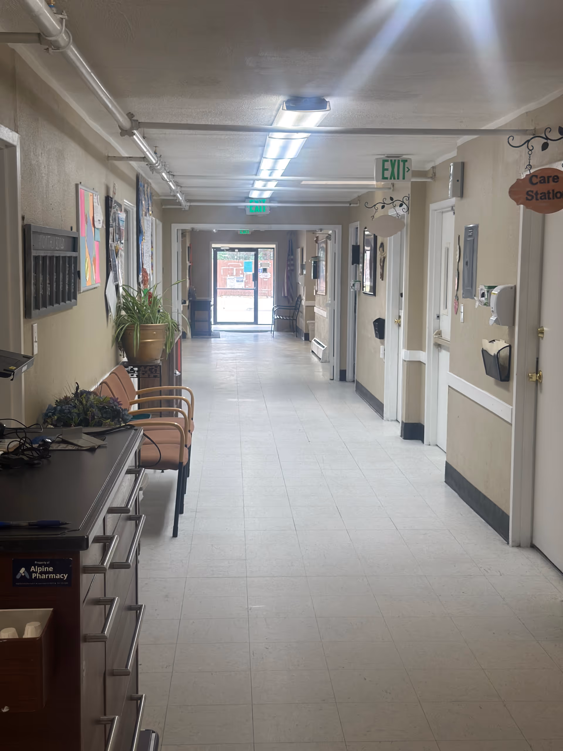 A long, well-lit hallway in an assisted living facility with beige walls and white tiled floor. There are chairs and plants along the left side, bulletin boards on the walls, and several doors on the right side. Exit signs are visible on the ceiling, and a door at the end of the hallway leads outside.