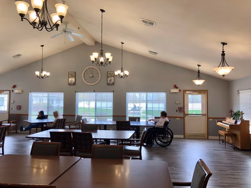 A spacious dining room with multiple tables and chairs arranged neatly. Three elderly individuals are seated, one in a wheelchair near a door with windows letting in natural light. The room has high ceilings with hanging chandeliers and ceiling fans. A piano with a plant on top is placed against the right wall. Large windows provide a view of the outdoor area.