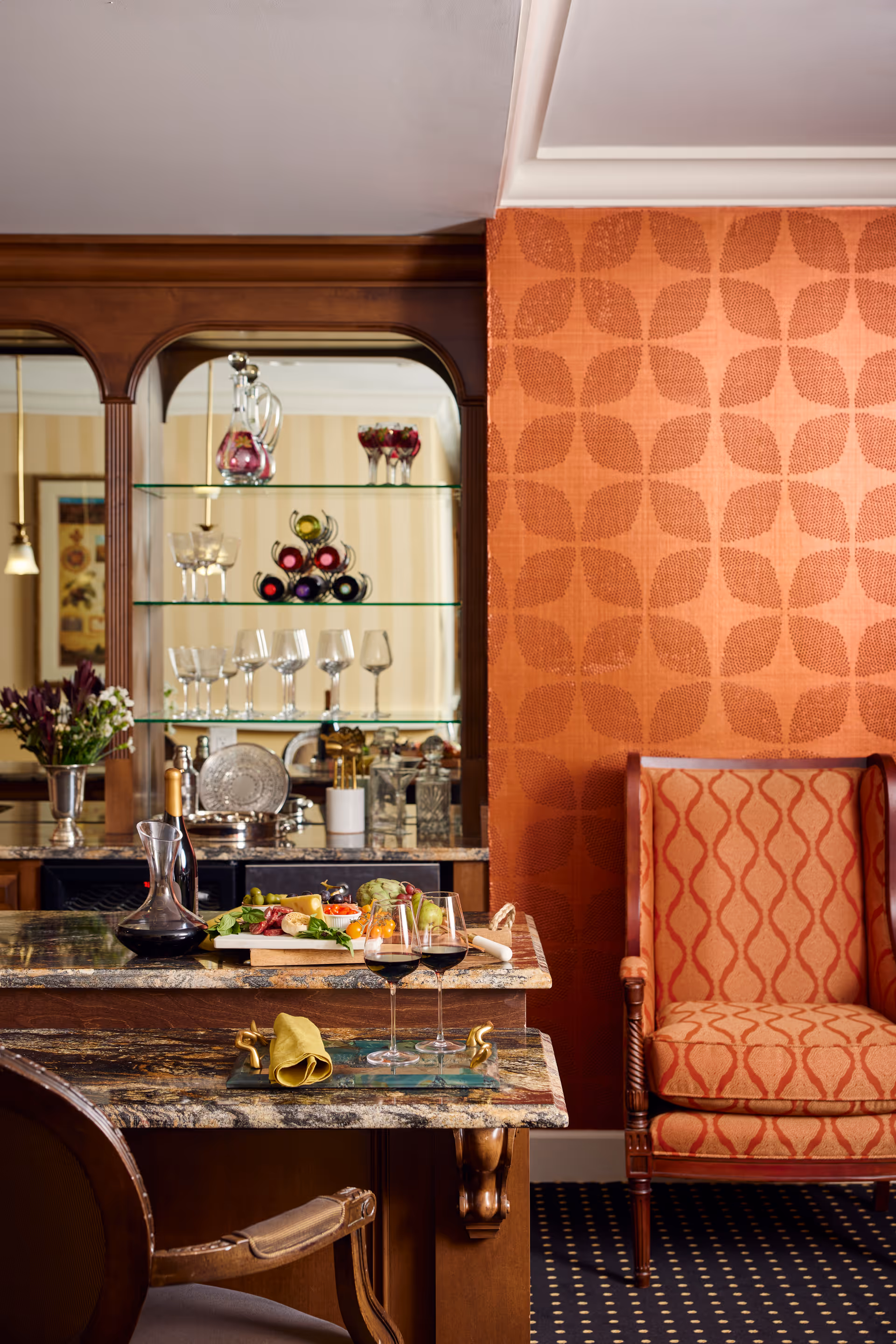 A cozy interior space featuring a marble countertop with two glasses of red wine, a decanter, and a charcuterie board with various foods. Behind the counter is a wooden cabinet with glass shelves displaying wine bottles and glassware. To the right, there is an upholstered armchair with a patterned orange fabric matching the orange patterned wallpaper behind it.