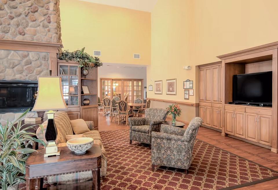A cozy living room area in a senior living facility with patterned armchairs, a sofa, a wooden side table with a lamp and decorative bowl, a stone fireplace, built-in wooden cabinets with a TV, and a dining area with chairs and tables visible in the background.