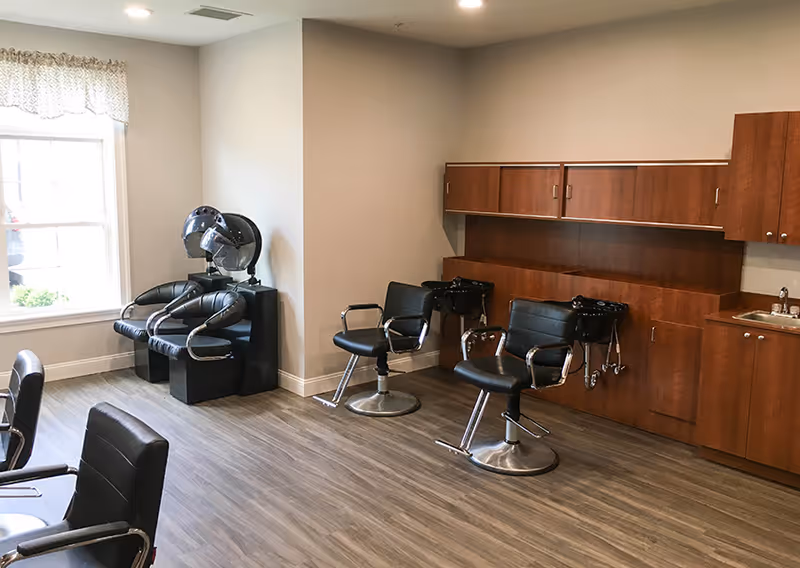 Interior of a salon area in a senior living facility with black salon chairs, hair washing stations, wooden cabinets, a sink, and a window with a valance.