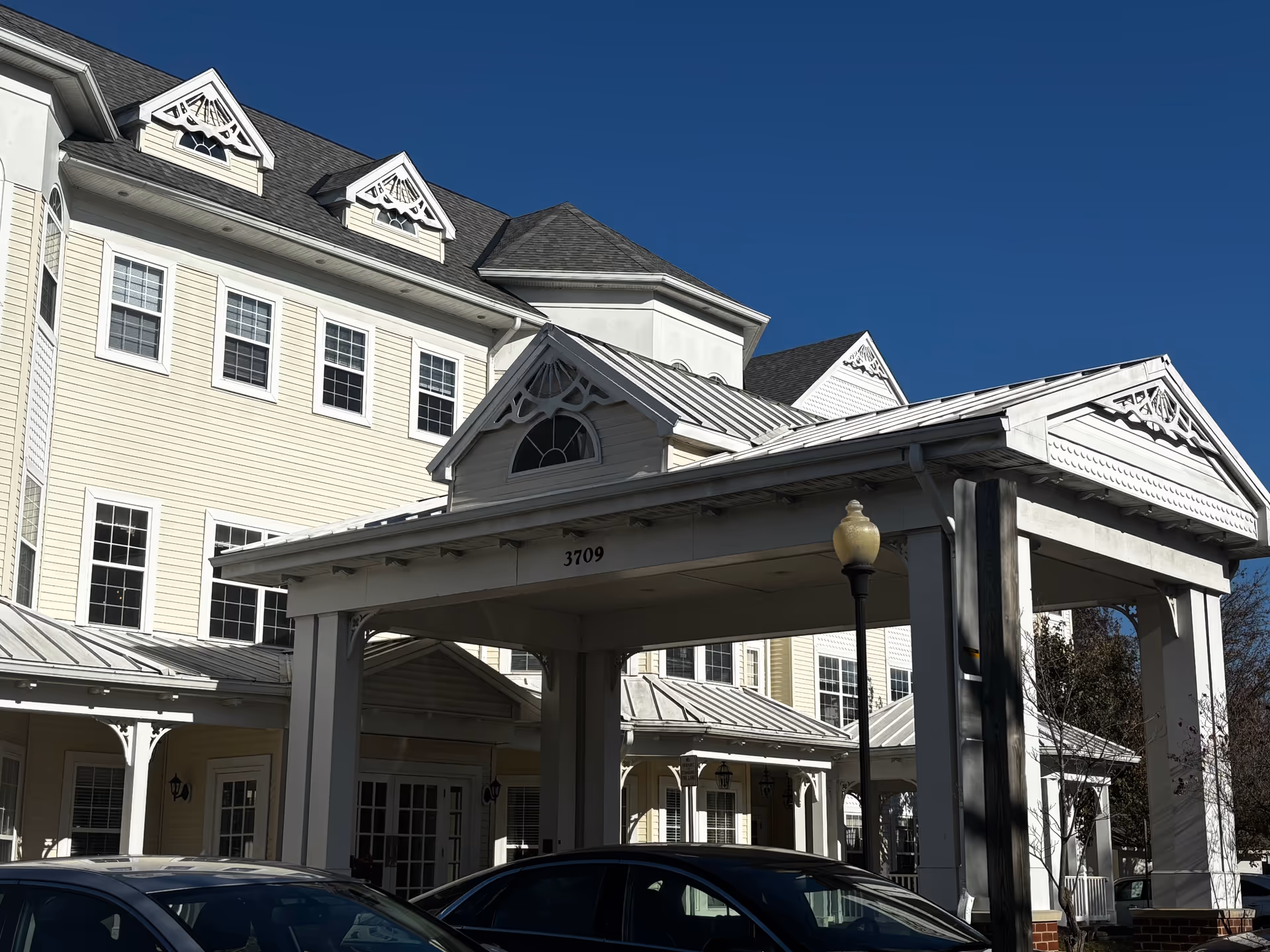 Exterior view of a senior living facility building with a covered entrance and multiple windows under a clear blue sky. Several cars are parked in front of the entrance.