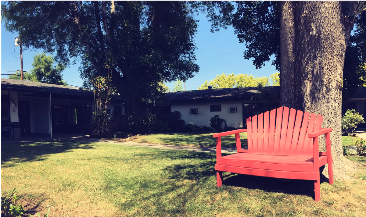 A bright red wooden bench with armrests is placed on a grassy lawn under a large tree. In the background, there is a single-story building with windows and air conditioning units, surrounded by shrubs and other trees under a clear blue sky.