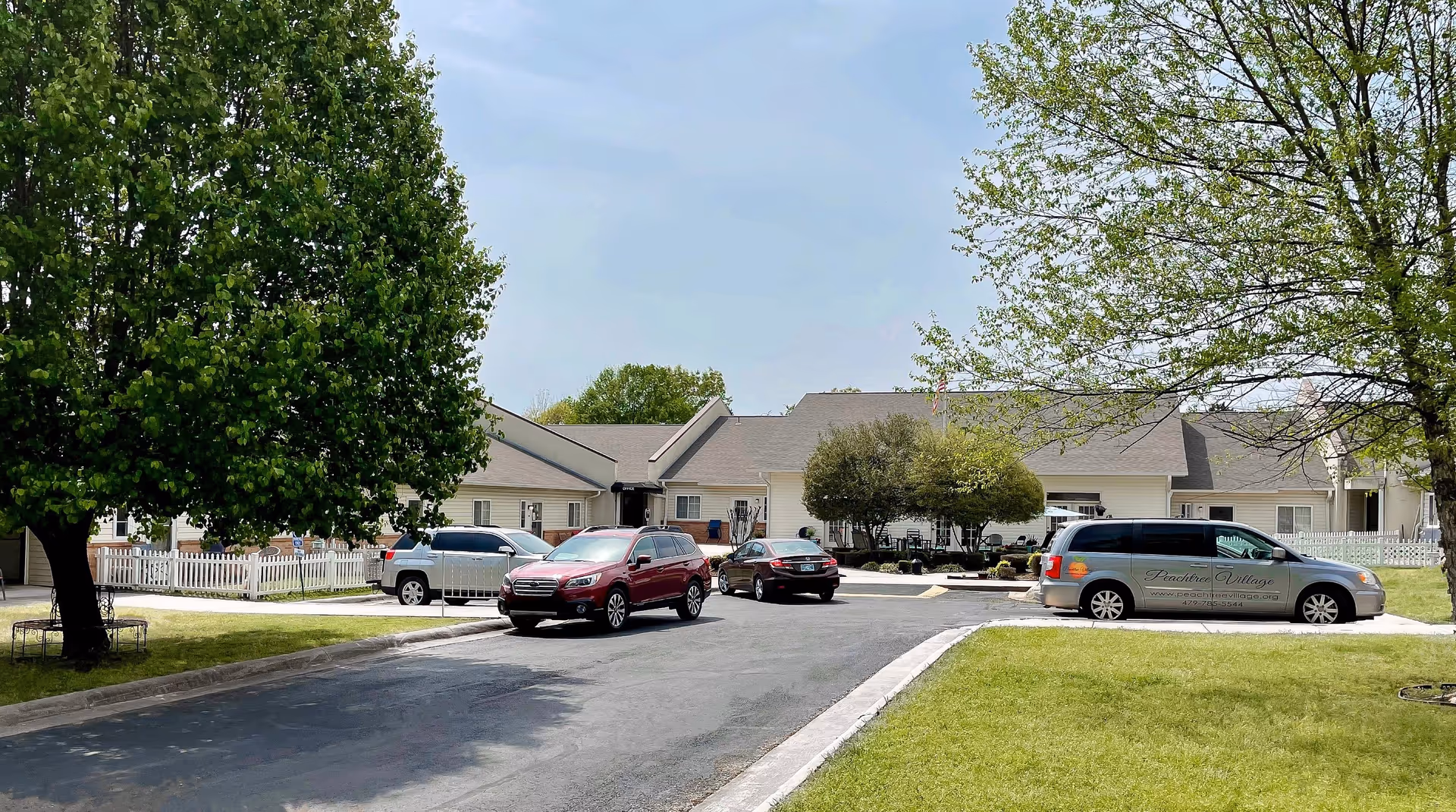Exterior view of Peachtree Village Assisted Living of Fort Smith showing a single-story building with a gray roof and light-colored siding. Several cars are parked in front of the building, including a van with the Peachtree Village logo and contact information. There are green trees and grass surrounding the parking area under a clear sky.