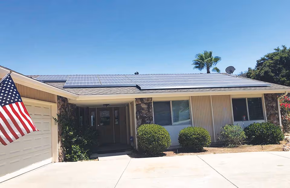 Front exterior of a single-story building with roof solar panels, an American flag, and shrubs along a driveway.