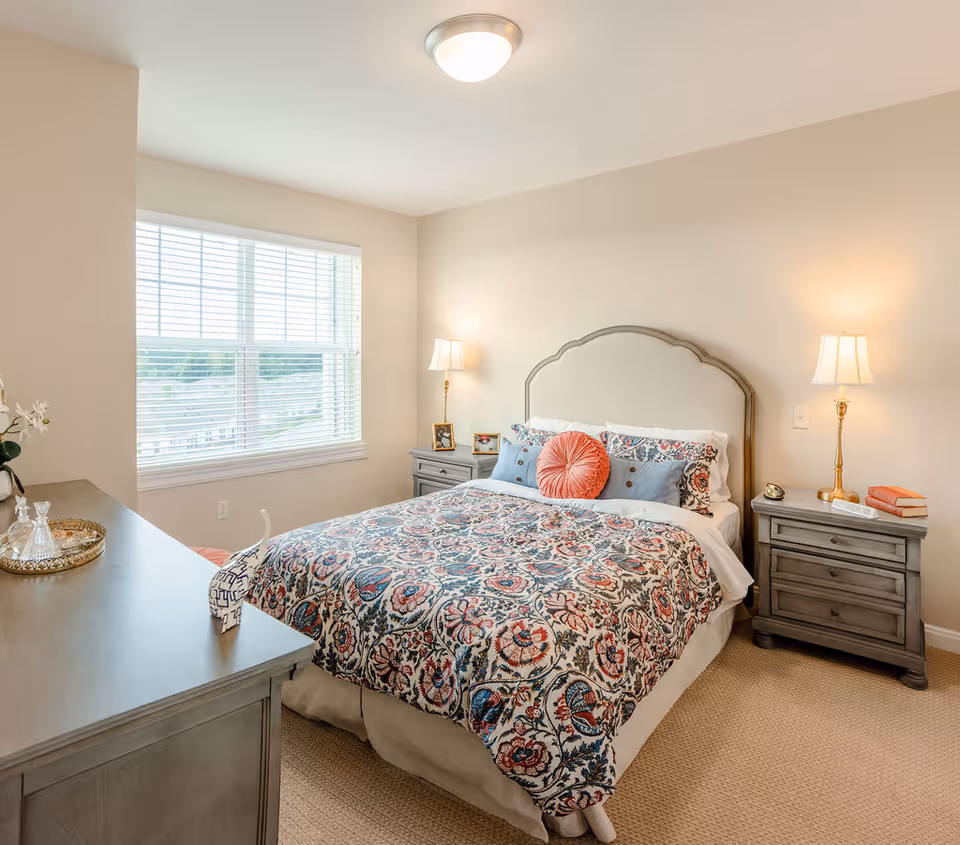A bright bedroom with a large window covered by white blinds. The room features a bed with a decorative floral-patterned bedspread, multiple pillows including a round coral-colored one, and a light gray upholstered headboard. On each side of the bed are matching gray nightstands with table lamps, picture frames, and books. A gray dresser with decorative items is positioned near the window.