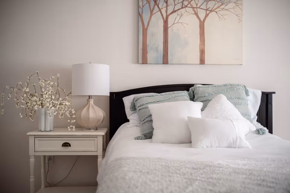 A neatly made bed with layered pillows, a nightstand holding a lamp and vase of flowers, and tree artwork on the wall above.