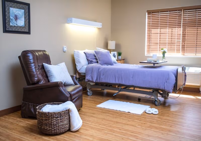 A cozy room with a hospital-style bed covered in purple bedding and pillows, a brown leather recliner chair with a white pillow, a wicker basket with a white blanket, a small white rug, slippers on the wooden floor, a window with wooden blinds, and a nightstand with a lamp and a small plant.