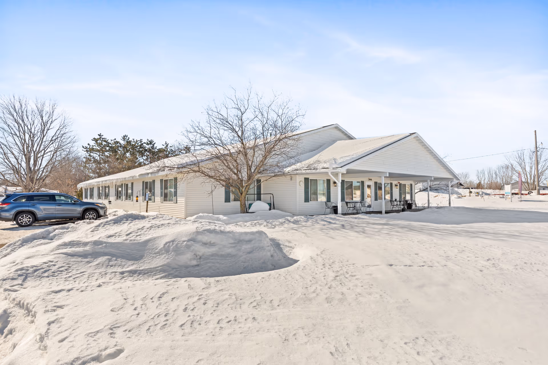 Exterior view of a single-story white building with a covered entrance and several windows, surrounded by snow-covered ground and bare trees under a clear blue sky. A parked car is visible on the left side.