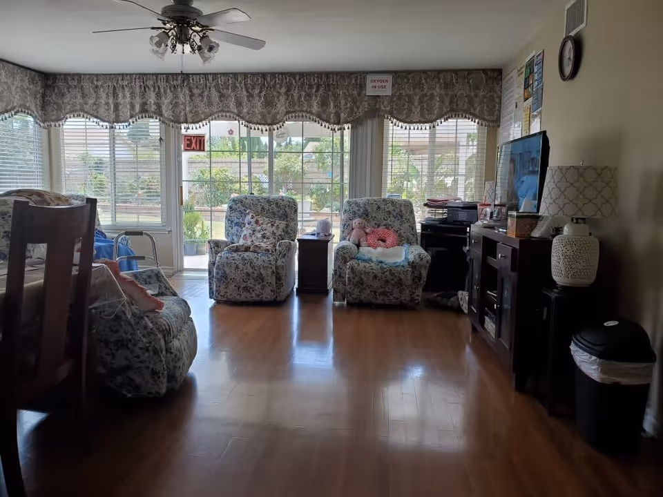 Sunlit common living room with floral recliners, hardwood floor, TV cabinet and large windows overlooking a garden.