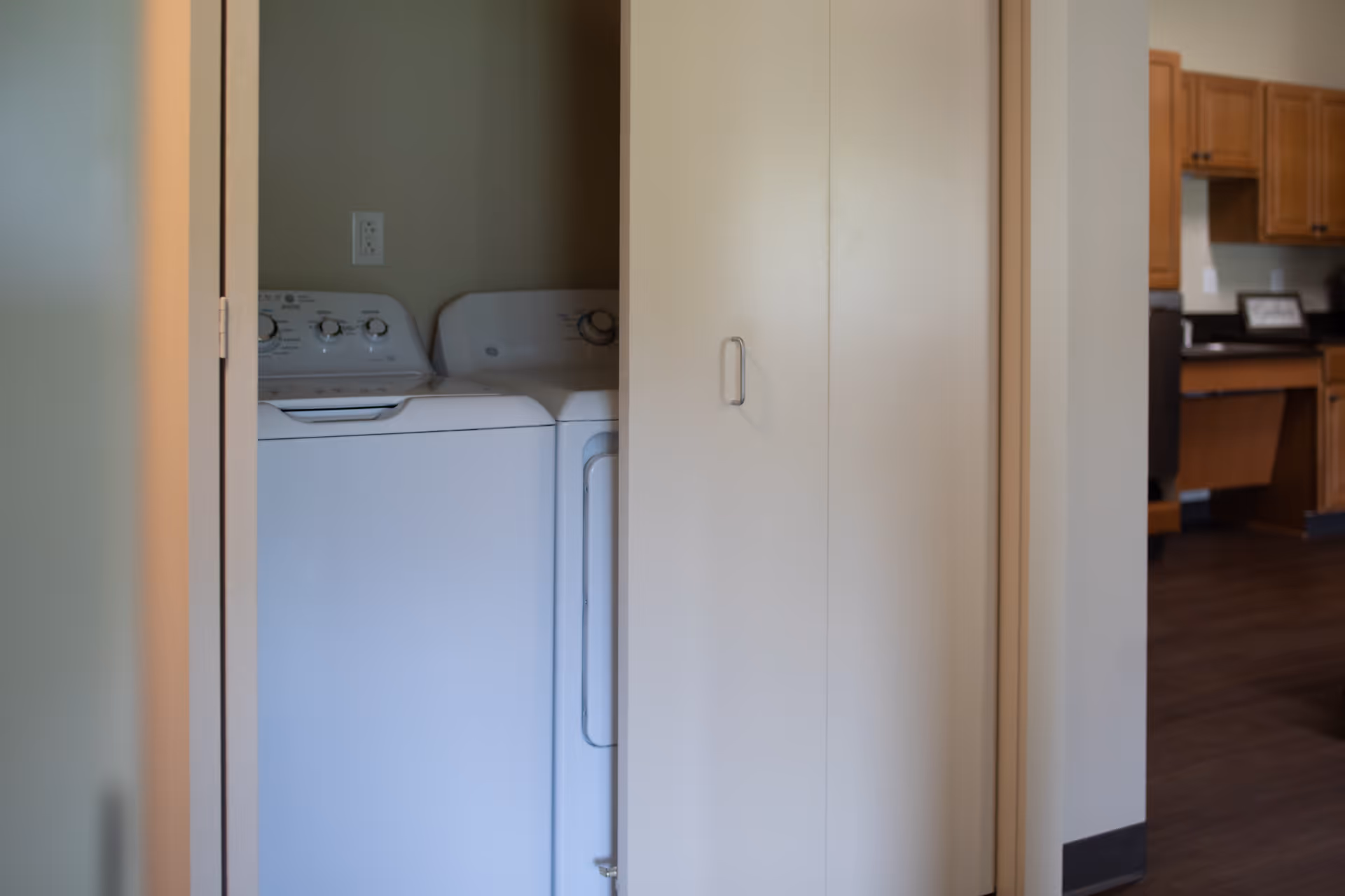 A laundry area with a white washing machine and dryer partially visible behind a partially open door, adjacent to a kitchen with wooden cabinets and dark countertops.