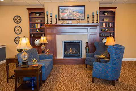 A cozy senior living common area with a lit fireplace centered on a wooden mantel. Two blue patterned armchairs and a matching loveseat are arranged around the fireplace. There are two side tables with lamps and decorative items. Built-in wooden shelves flank the fireplace, holding books and decorative pieces. A framed landscape painting hangs above the mantel.