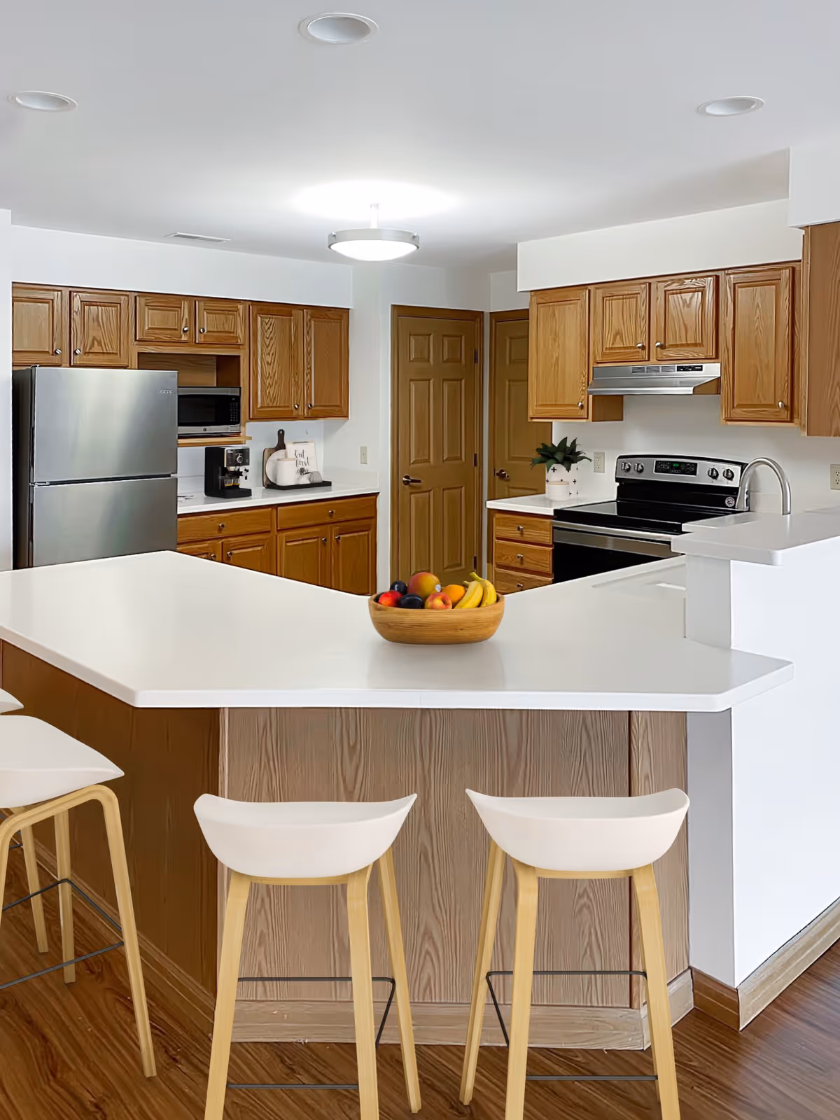 A modern kitchen with wooden cabinets, a stainless steel refrigerator, microwave, and stove. There is a white countertop island with four white bar stools and a wooden bowl filled with fruit on the island. The floor is wood, and there are two closed wooden doors in the background.