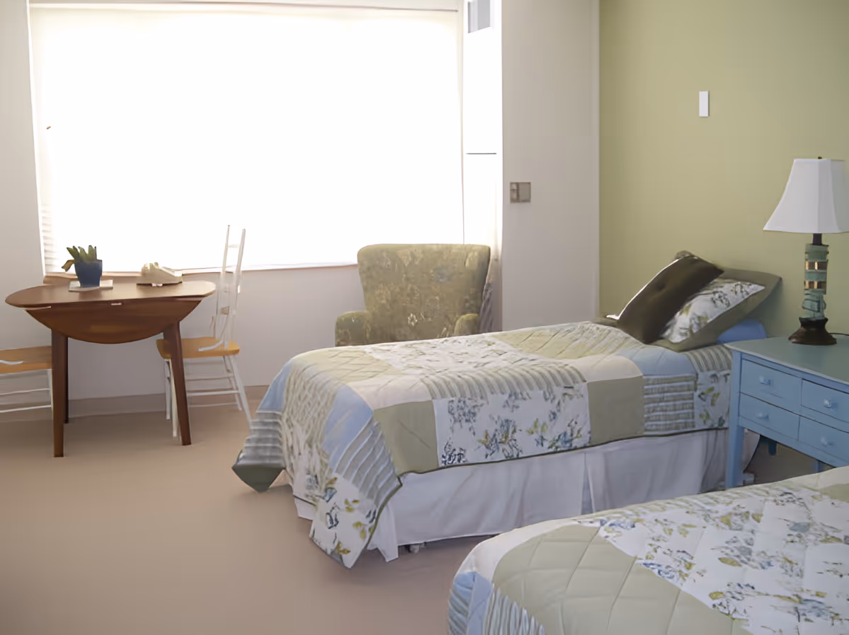 Sunlit bedroom with two twin beds covered in patchwork quilts, a blue bedside table and lamp, an armchair, and a small table by the window.